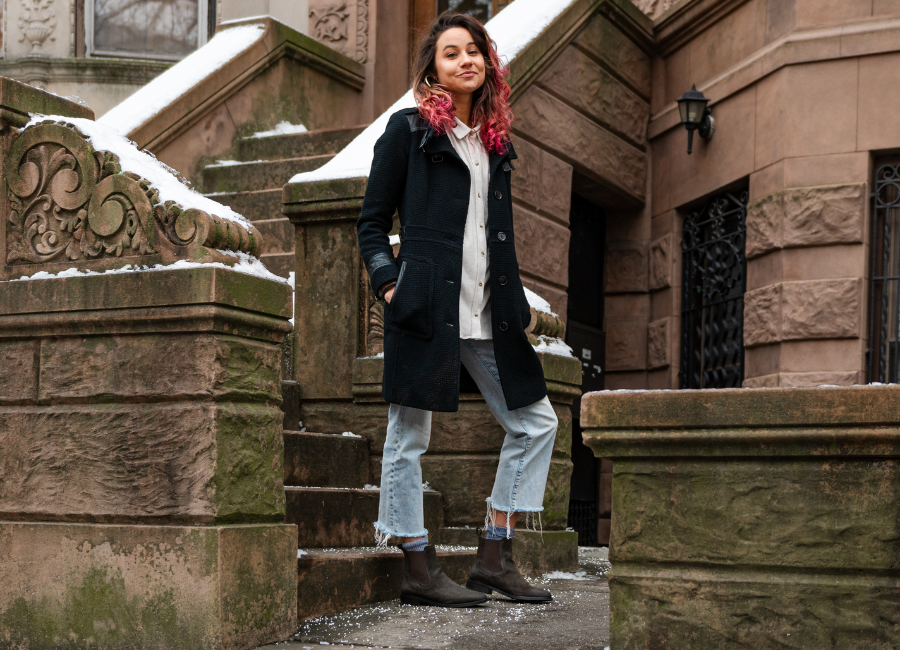 A women with pink highlights, standing in front of red stone stairs wearing a black coat, white blouse, stonewashed light blue denim jeans, and Thursday's Duchess chelsea boots in Green Olive suede